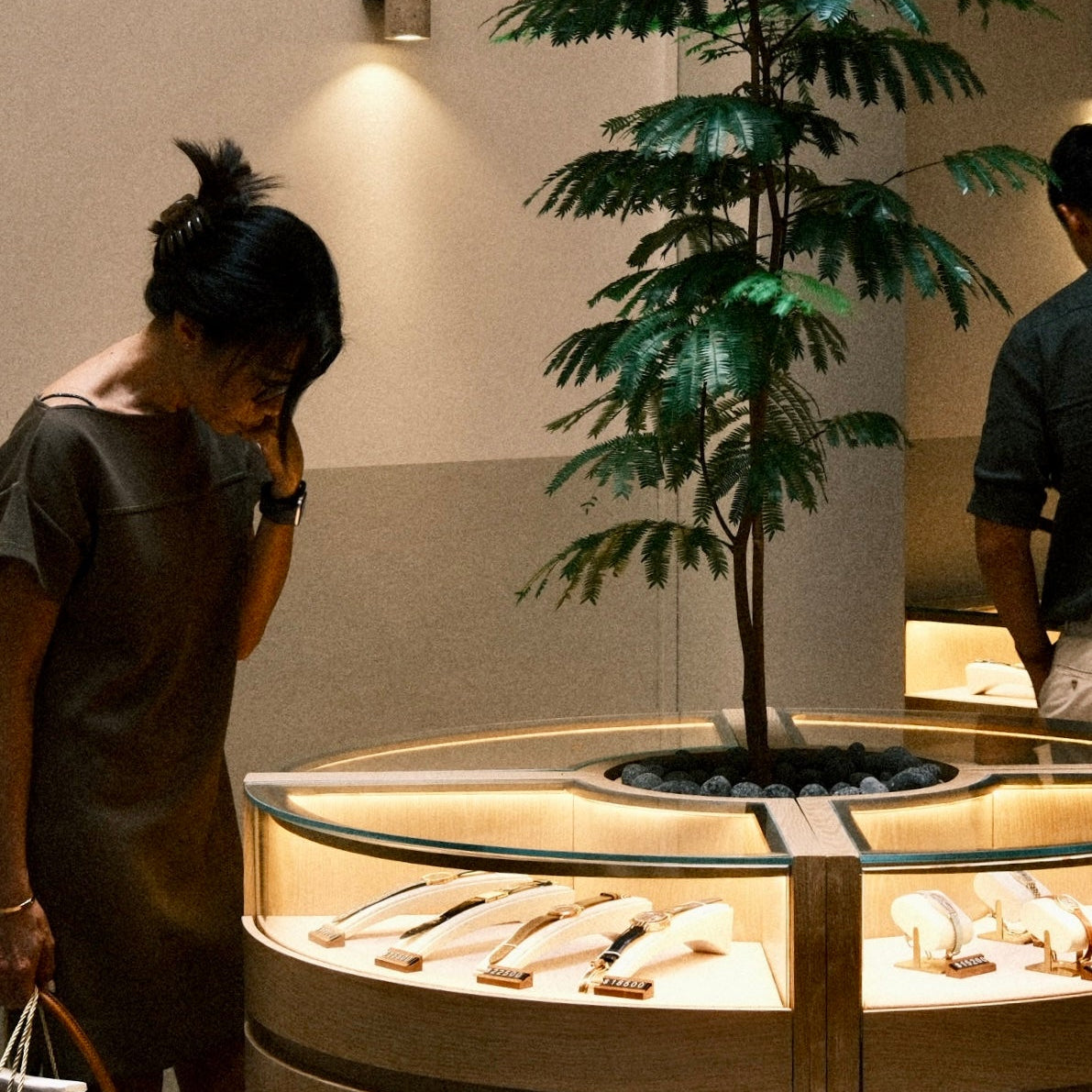 A woman in a brown dress looks down at a curved glass jewelry display case inside Reference Room. A tall potted green tree stands behind the case, and another person is partially visible on the right.