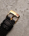 Close-up of the original 18k yellow gold Corum buckle, engraved with the Corum key logo, attached to the black textured leather strap.