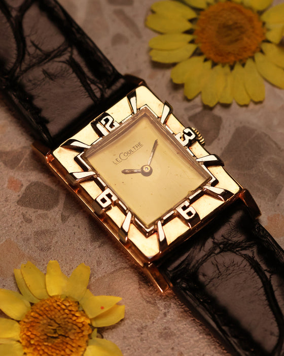 LeCoultre 14K gold tank watch placed on marble with yellow flowers, showcasing the interplay of gold tones and Art Deco numerals bezel.