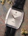 Rolex Cellini Ref. 4087 in 18K white gold with silver sunburst dial and black leather strap, photographed flat lay on a textured stone surface with soft florals.
