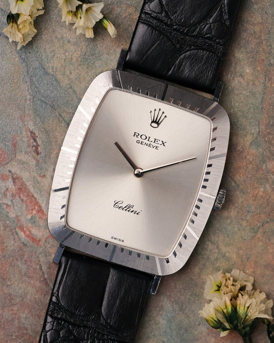 Rolex Cellini Ref. 4087 in 18K white gold with silver sunburst dial and black leather strap, photographed flat lay on a textured stone surface with soft florals.