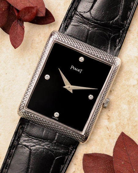 Piaget Ref. 9152 in 18K white gold with woven textured case, black onyx dial and diamond markers, photographed flat lay with black leather strap and red leaves.