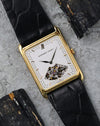 Audemars Piguet rectangular 18K yellow gold watch with white partially openworked dial photographed on a textured grey surface.