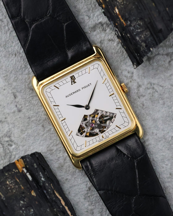 Audemars Piguet rectangular 18K yellow gold watch with white partially openworked dial photographed on a textured grey surface.