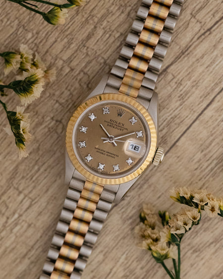 Rolex Tridor Datejust photographed with flowers, showing its champagne diamond dial and three-colour gold bracelet.