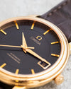 Macro view of the black Omega De Ville dial showing the applied Omega emblem, gold hands, markers and framed date aperture.