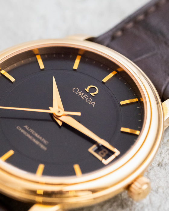 Macro view of the black Omega De Ville dial showing the applied Omega emblem, gold hands, markers and framed date aperture.