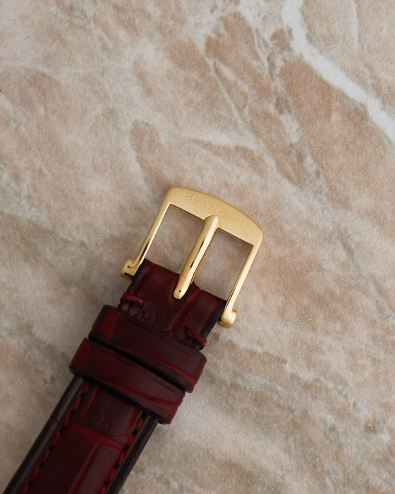 Front view of the Blancpain-signed yellow gold buckle fitted to a burgundy leather watch strap.