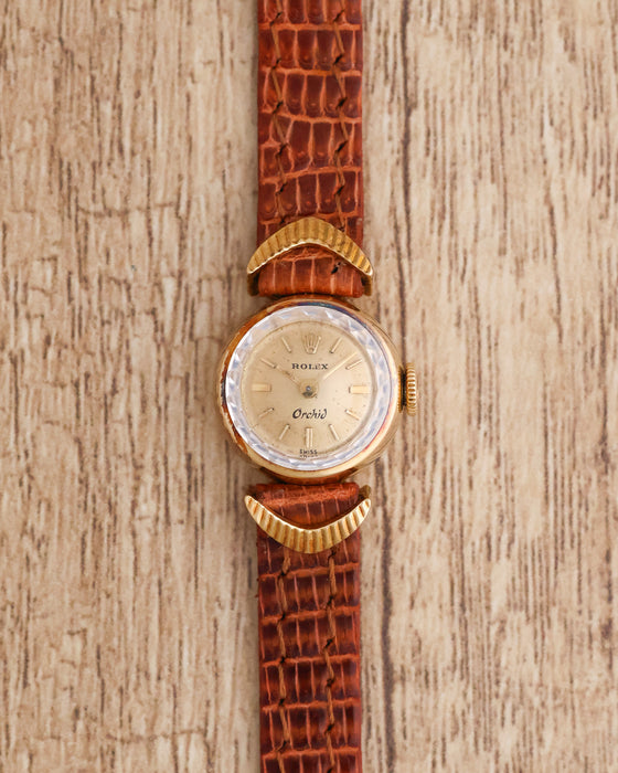 Front-facing view of a vintage Rolex Orchid Ref. 2132 in yellow gold with a champagne dial, sculpted lugs and brown leather strap.