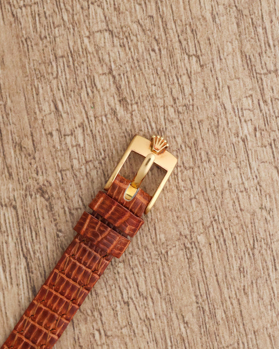 Close-up of the Rolex-signed yellow gold-tone pin buckle attached to a brown textured leather strap.